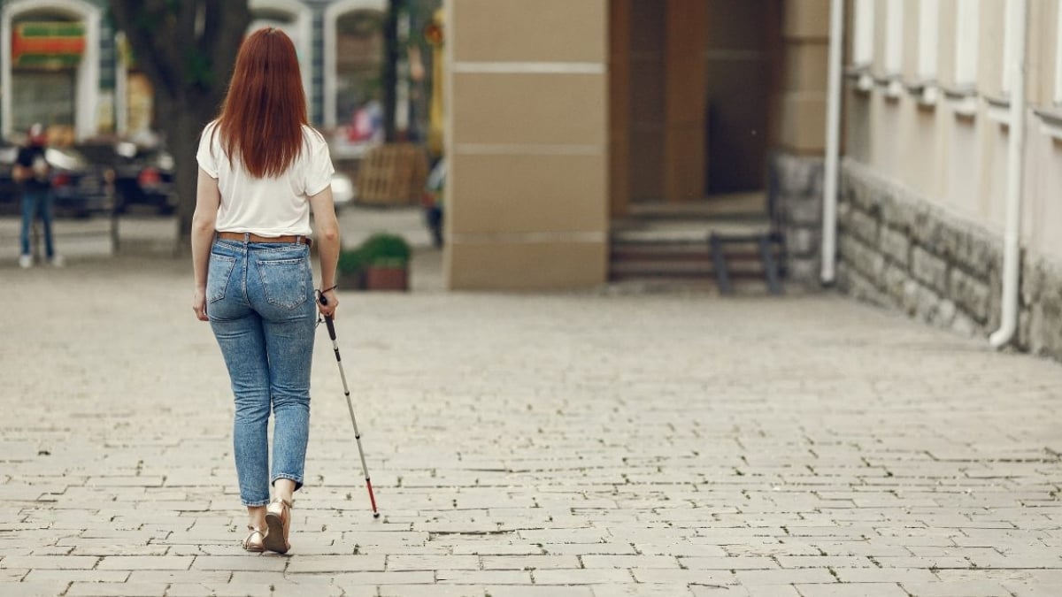 Fotografía de una mujer joven de cabello rojizo, que lleva puesto un pantalón de mezclilla azul y una blusa color beige; se encuentra caminando por una acera, va apoyada por un bastón guía para personas con discapacidad visual.