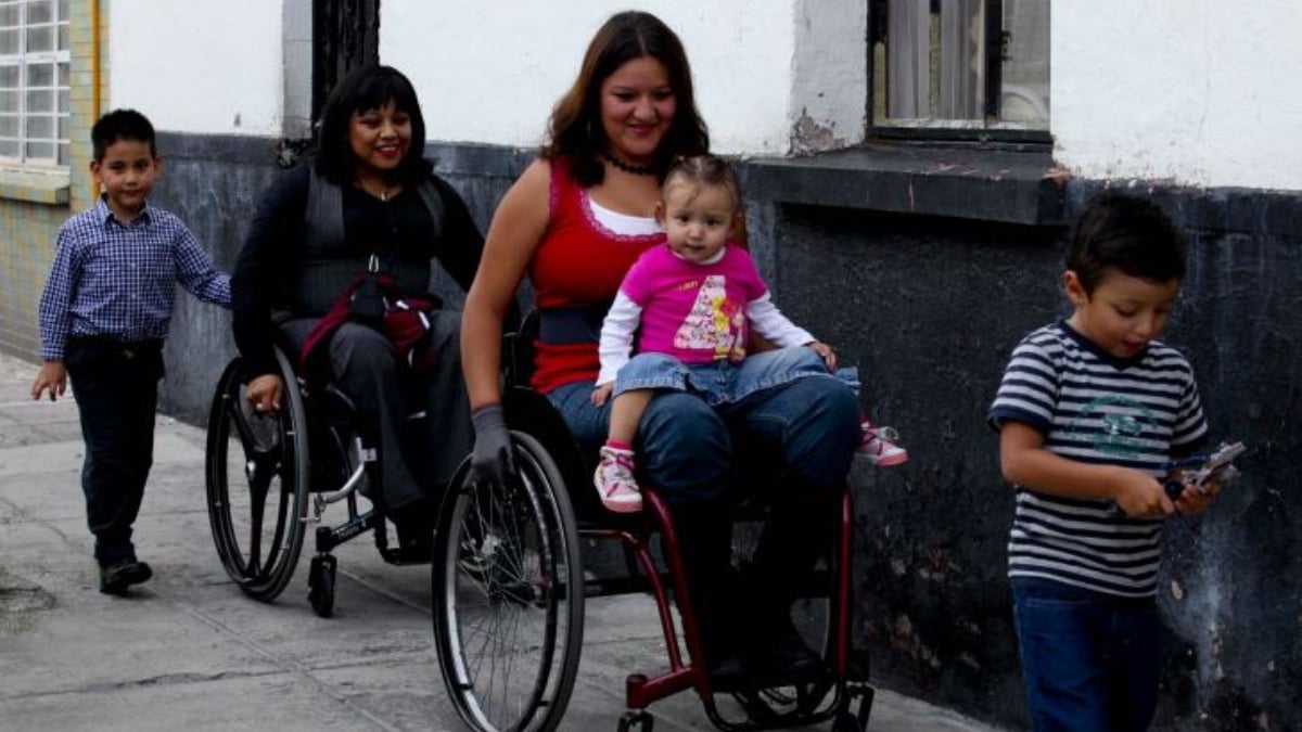 Fotografía de dos mujeres con discapacidad motriz usuarias de silla de ruedas que van por una banqueta formando una fila con tres menores. El primero es un niño que apenas se alcanza a ver, le sigue una mujer joven de cabello oscuro a la altura de los hombros, con una camiseta blanca sin mangan y encima otra roja, también sin mangas, y pantalón de mezclilla azul, sobre sus piernas lleva a una bebé de unos dos años con faldita azul y una playera rosa con un estampado al frente; le sigue otra mujer con su silla de ruedas, que viste ropa oscura y tiene el caballo negro, y al final camina otro niño de unos cinco años, que viste pantalón oscuro y playera estampada en tonos grises.