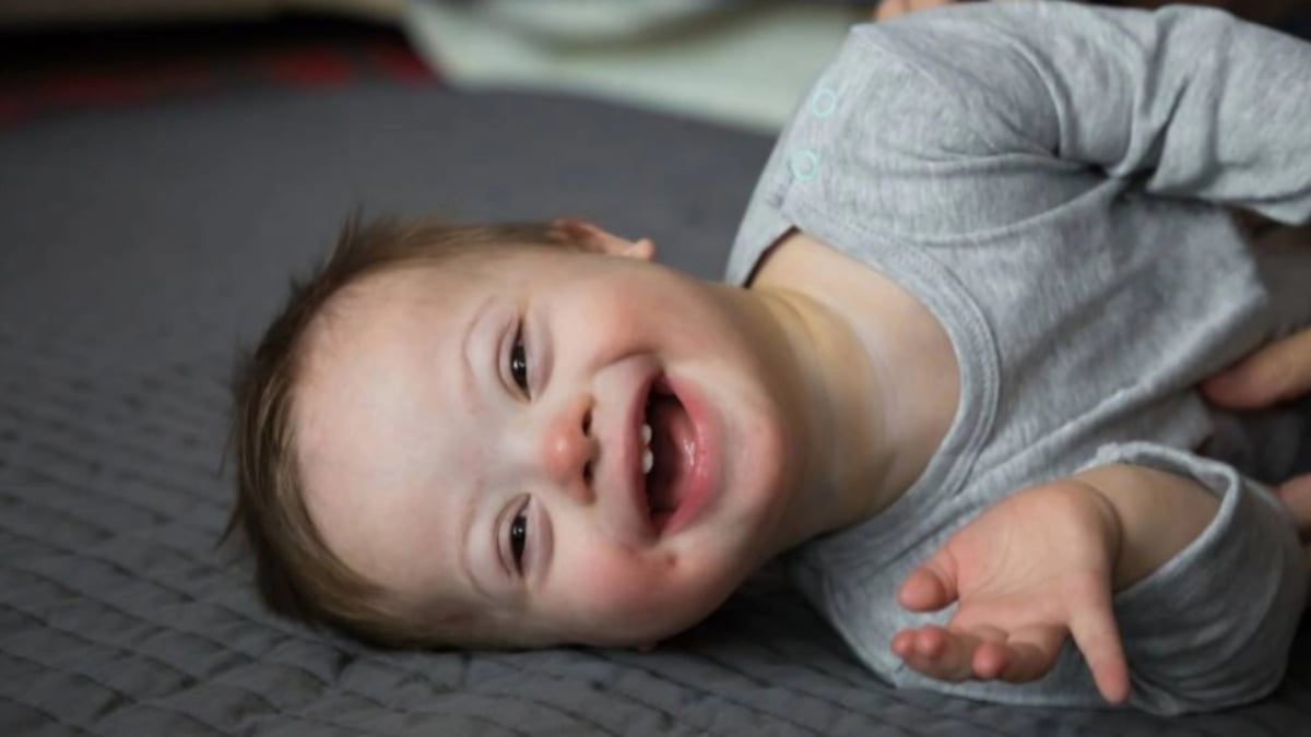 Fotografía de un bebé de menos de un año que tiene síndrome de Down, está recostado sobre su lado izquierdo sobre una superficie acolchada color gris de tal manera que la fotografía se ve de lado. El bebé tiene tez blanca, cabello rubio y una enorme sonrisa. Estira su brazo izquierdo y usa una especie de camiseta gris de manga larga.