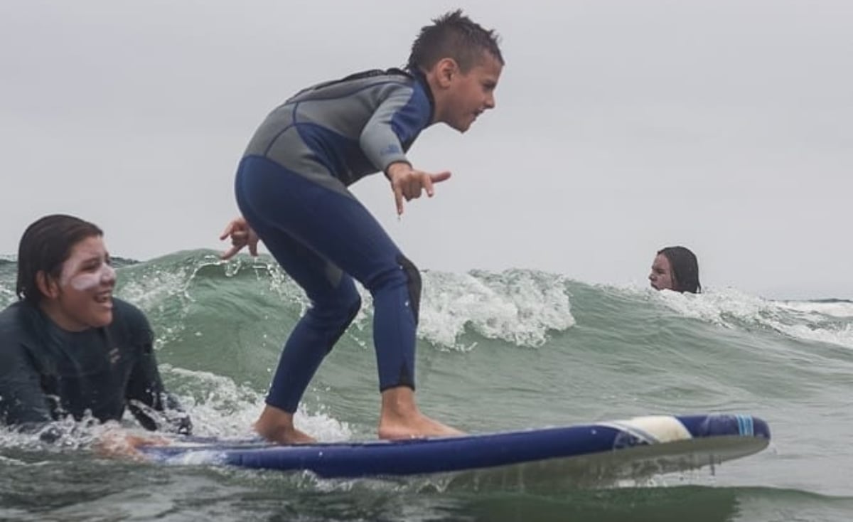 Fotografía de un niño como de 10 años arriba de una tabla de surf dentro del mar, abre sus brazos para equilibrarse mientras llega una ola que está solo un par de pasos detrás de él. El niño es supervisado por dos personas adultos, una muy cerca del final de la tabla y la otra detrás de la ola. El niño viste un traje completo de surfer en colores azul y gris.