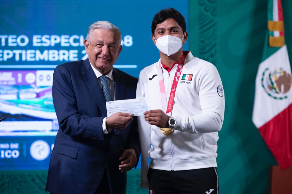 Fotografía horizontal de Juan Diego García, atleta paralímpico de cabello castaño oscuro que lleva puesto un cubrebocas KN95 y viste una chamarra deportiva de color blanco, pants deportivo de color negro y tenis de color blanco, se encuentra de pie a lado del presidente de México, Andrés Manuel López Obrador, un hombre de edad adulta, cabello cano, tez morena que viste un traje formal de color azul, corbata plateada, calza zapatos de color negro y entrega con su mano derecha un cheque a Juan Diego, quien a su vez toma el cheque con su mano derecha, ambos sonríen frente a la cámara.