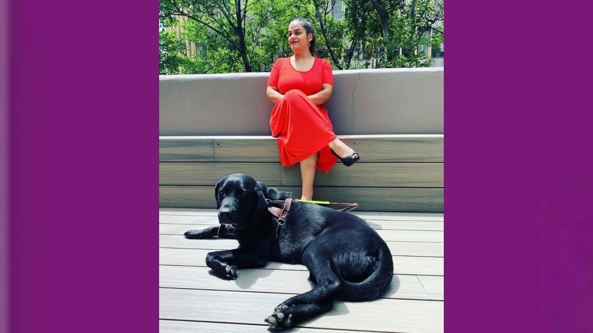 Fotografía de Lucero Márquez, una mujer de cabello oscuro, recogido, que usa un vestido largo, rojo, y está sentada en una banca con la pierna cruzada mientras sonríe, frente a ella está Kyler, su perra guía, es una labrador negra de pelo brillante, que está acostada con la cabeza erguida.