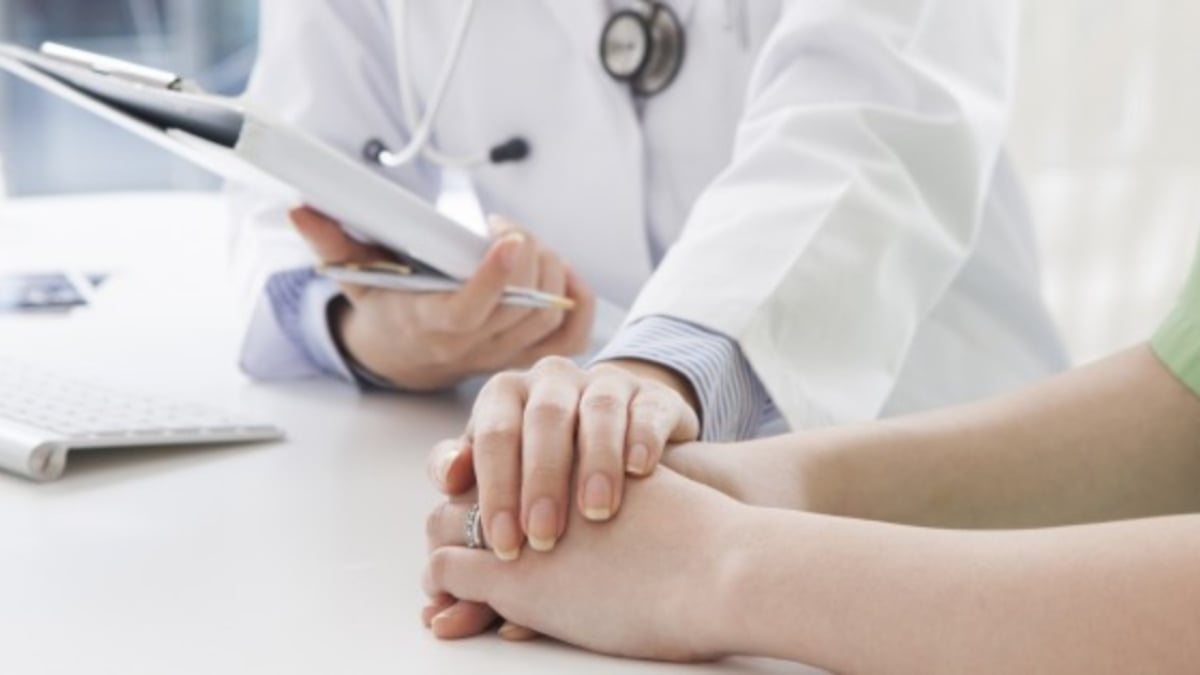 Fotografía en la que se ven las manos de dos personas. Una de ellas está vestida con una bata de médico, sobre una blusa azul, tiene un estetoscopio colgando y una tableta en la mano derecha mientras coloca la izquierda sobre las dos manos de otra persona.