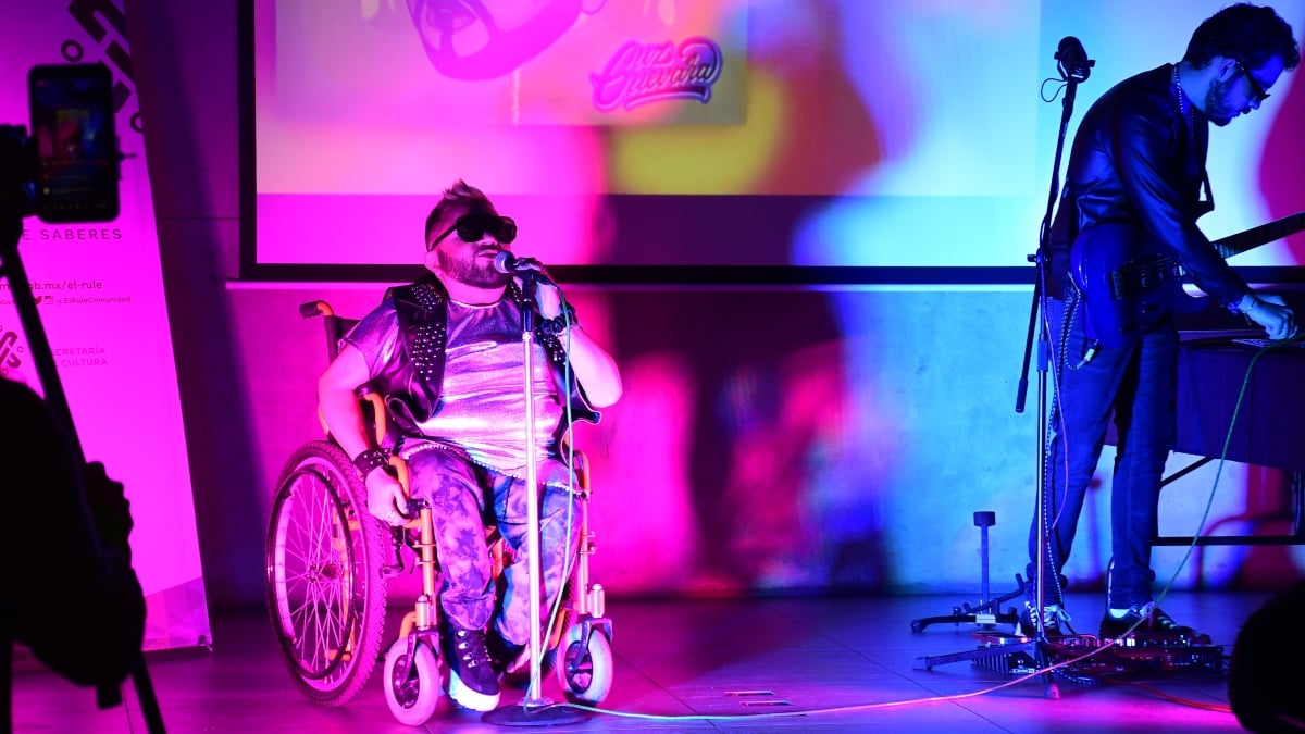 Fotografía de un hombre joven usuario de silla de ruedas a mitad de un escenario iluminado con luces rosas y azules. El hombre tiene un micrófono en la mano izquierda, pantalón y camiseta estampados y chaleco con estoperoles. A un lado de él, de pie, está un hombre frente a un teclado.