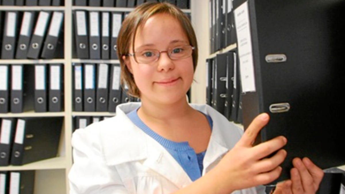 Fotografía de una adolescente con síndrome de Down, de tez blanca, con cabello rubio corto, con lentes cuadrados, vestido azul y encima un bata blanca que guarda una carpeta negra en un archivero; está dentro de un cuarto para archivo.