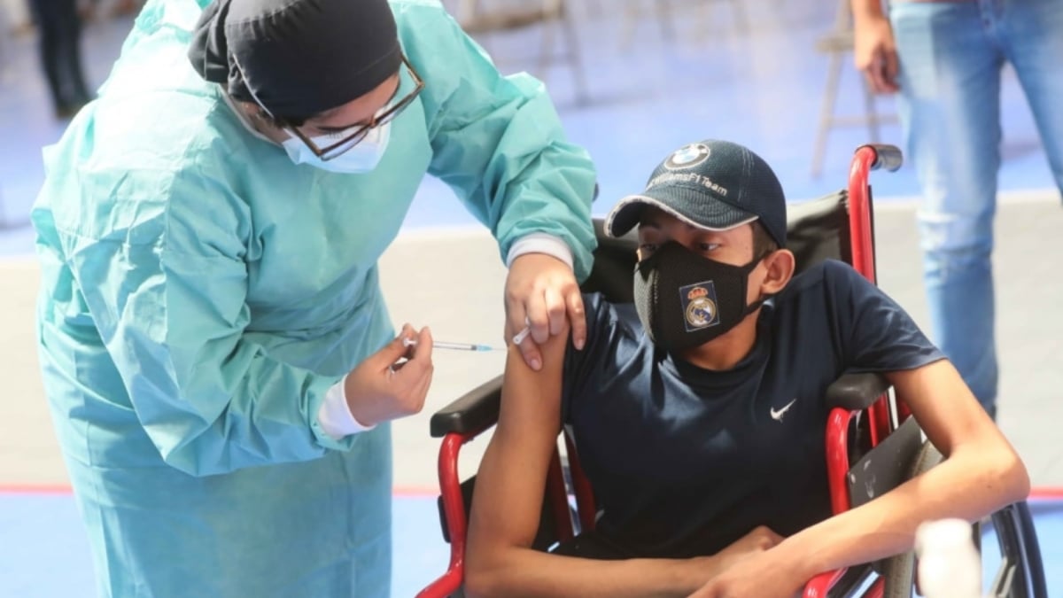 Fotografía de un adolescente usuario de silla de ruedas, que viste camiseta deportiva color negro, cubrebocas y gorro del mismo color,cuando recibe una vacuna en el brazo derecho, que le es aplicada por una mujer enfermera, que viste traje estéril color azul agua, con cofia negra y cubrebocas blanco.