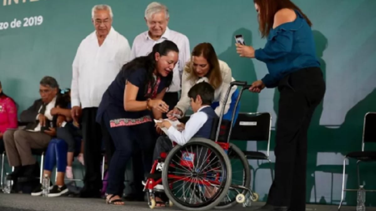 Fotografía de un templete usado en un acto oficial de gobierno en la que aparece un grupo de personas, algunas con discapacidad. El primer plano de la imagen son dos mujeres, semi inclinadas, que entregan una tarjeta bancaria a una persona usuaria de silla de ruedas, mientras dos hombres adultos mayores, con cabello canoso y vistiendo guayaberas color blanco, las ven y sonríen. A la derecha, hay una mujer de pie, que viste pantalón negro y blusa azul que tiene un celular en la mano izquierda y captura la escena.