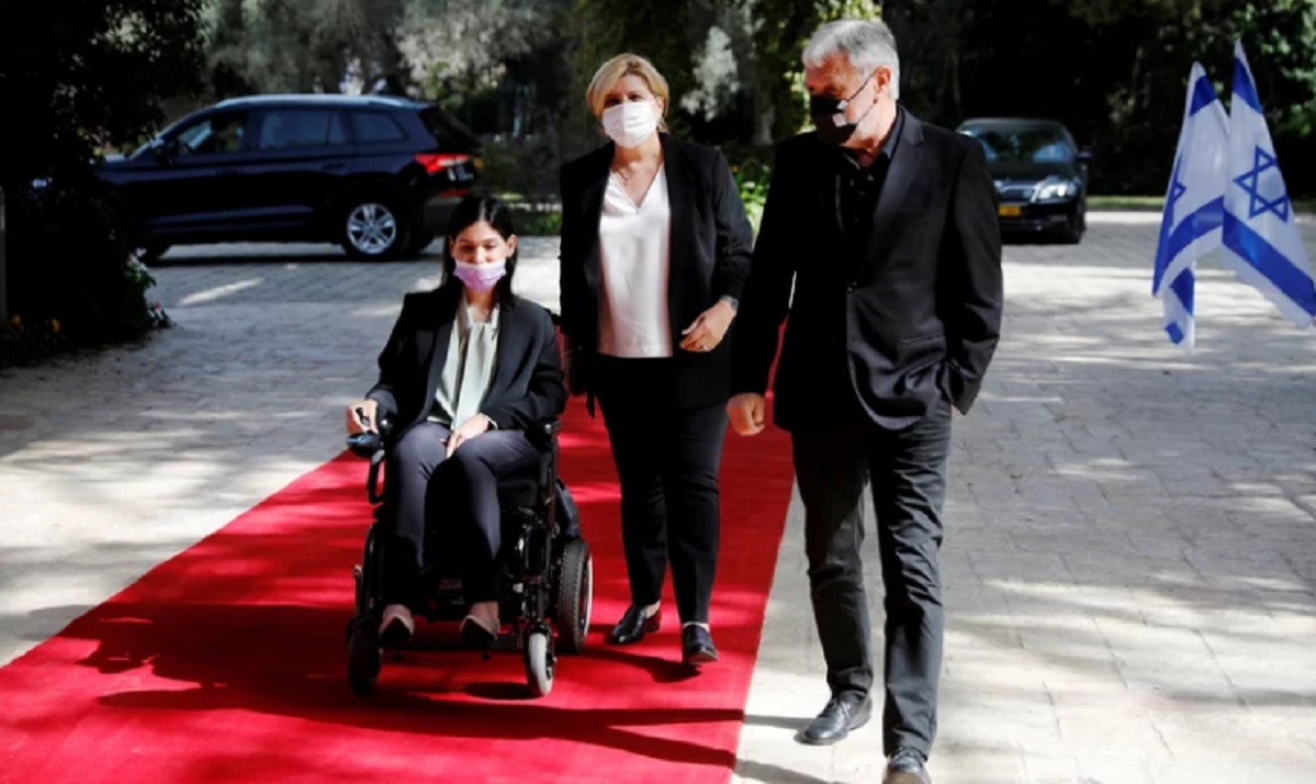 Fotografía de archivo, de abril de 2021, en la que se ve una alfombra roja con tres personas. Una de ellas es una mujer joven, de cabello oscuro, usuaria de silla de ruedas. Viste traje negro, blusa gris y un cubrebocas gris. A su lado, de pie, una mujer de cabello rubio, que usa cubrebocas blanco, blusa blanca y saco y pantalón negro, y un hombre también de pie, de cabello canoso y vistiendo riguroso negro. A la derecha del hombre se ven dos banderas del Estado de Israel.