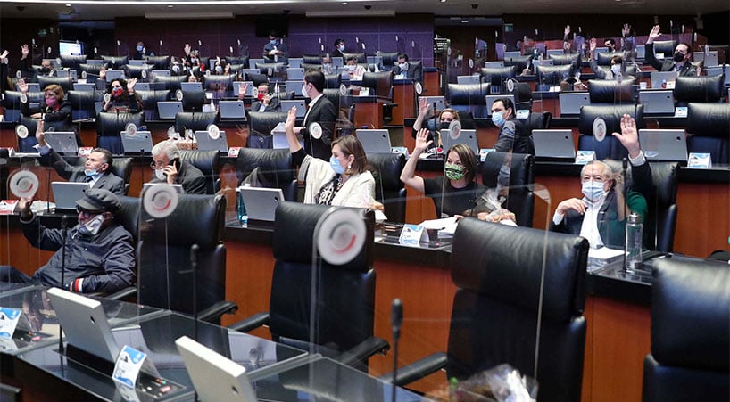 Fotografía una parte de la sala del Senado de la República, sillas de color negro, acolchonadas, divididas por un muro de plástico entre cada lugar, en la fotografía se alcanzan a apreciar siete pisos de escritorio, silla y computadoras, en ella se ven hombres y mujeres con la mano levantada ejerciendo su voto, todos con cubrebocas.