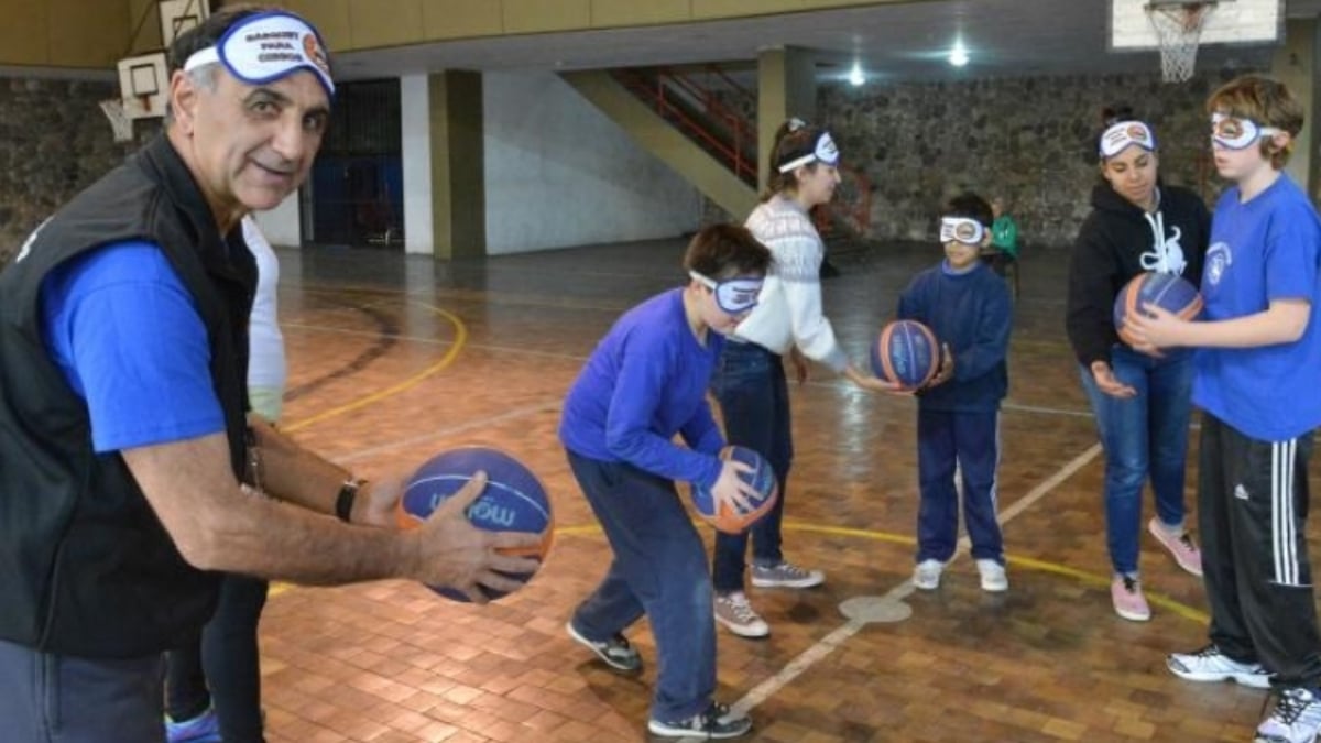 Fotografía de un hombre adulto mayor que tiene en las manos un balón azul de basquetbol y entrena a un grupo de niños y adolescentes con discapacidad que también tienen su respectivo balón, mientras están en una cancha de ese deporte. Todos visten pants azul oscuro y camisa azul rey.