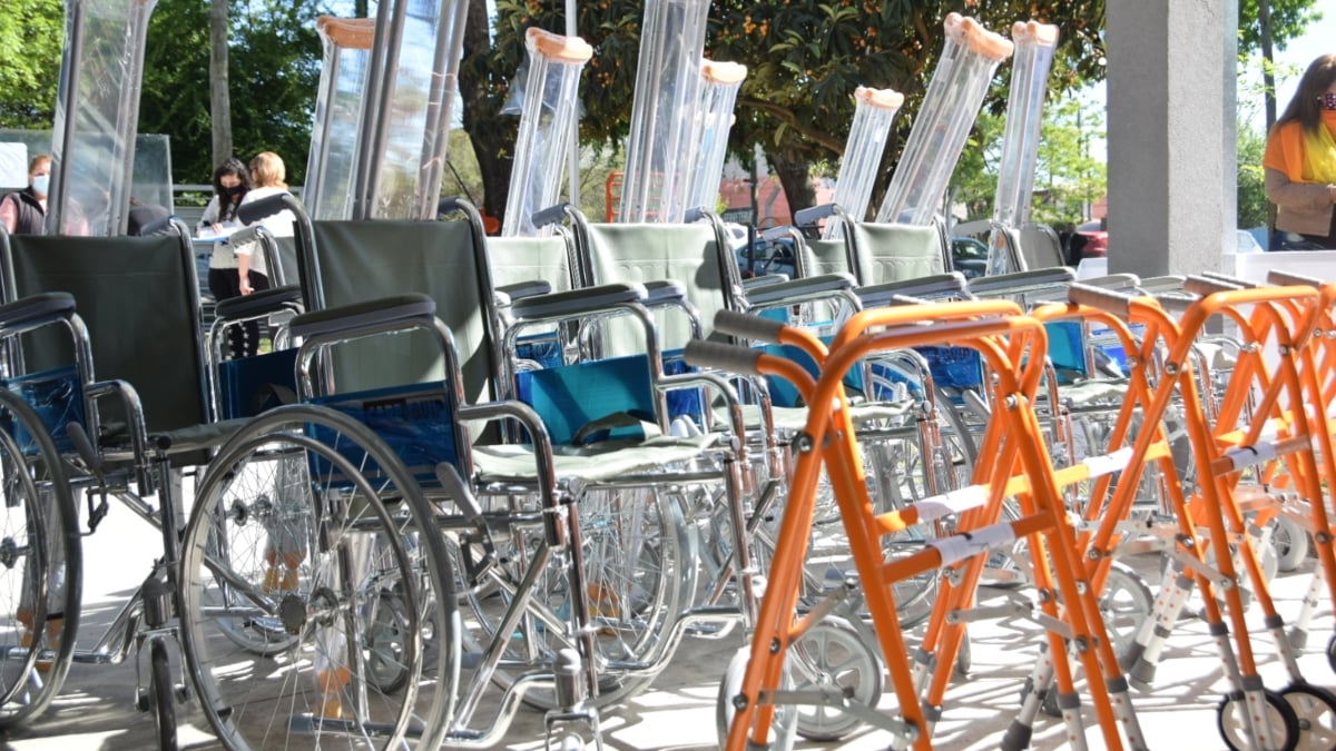 Fotografía de un grupo de sillas de rueda vacías, frente a ellas un grupo de andaderas color anaranjado y, atrás de las sillas, varios pares de muletas empacados.