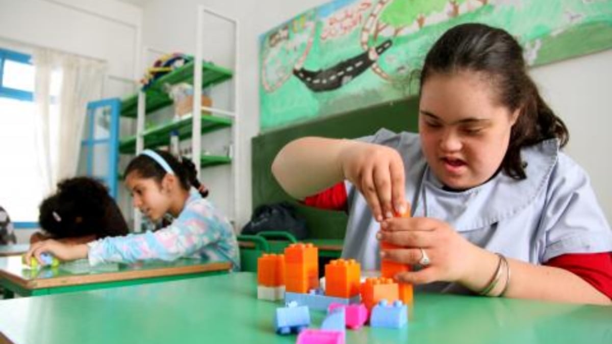 Fotografía de UNICEF en la que se ve a tres niñas, dos al fondo y una en primer plano dominando la imagen está una adolescente con discapacidad, que juega con unos bloques de colores; ella tiene la tez blanca y cabello oscuro peinado en media cola.