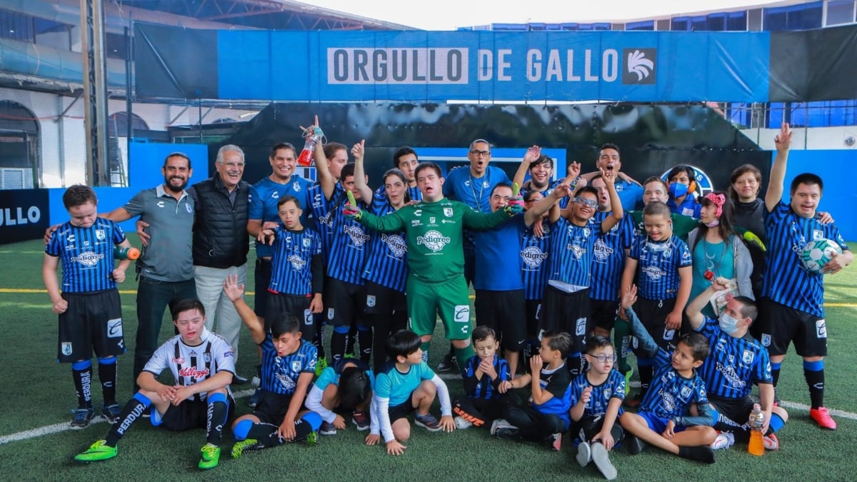 Fotografía del equipo de fútbol inclusivo Gallos-Smiling, conformado por personas con discapacidad intelectual. Los jugadores están colocados en varias filas desordenadas, que incluyen a algunos miembros en cuclillas al frente de todos. Los jugadores visten uniformes azules y se colocan frente a un letrero azul que dice Orgullo de Gallo