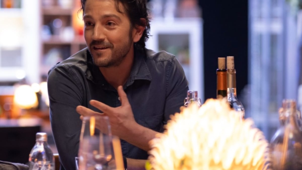 Fotografía de un hombre joven, de tez blanca, cabello oscuro, corte desigual, barba corta y bigote, viste una camisa gris oscura, está sentado frente a una mesa, a su lado hay un par de botellas con corcho, frente a él una copa y un arreglo floral. Voltea un poco la cara al lado derecho y sonríe, mientras coloca la mano izquierda frente a él y la abre.