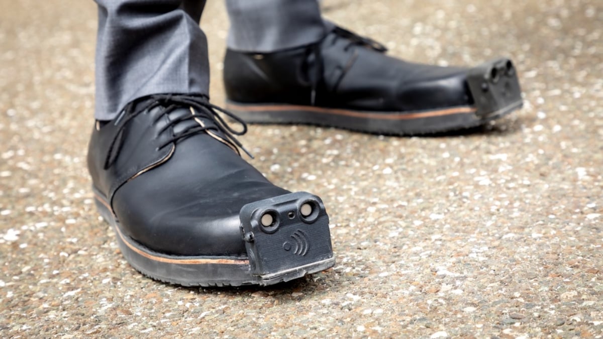 Fotografía que muestra un par de zapatos negros que en la punta tienen colocado un sensor cuadrado que ofrece información del entorno a las personas con discapacidad visual.