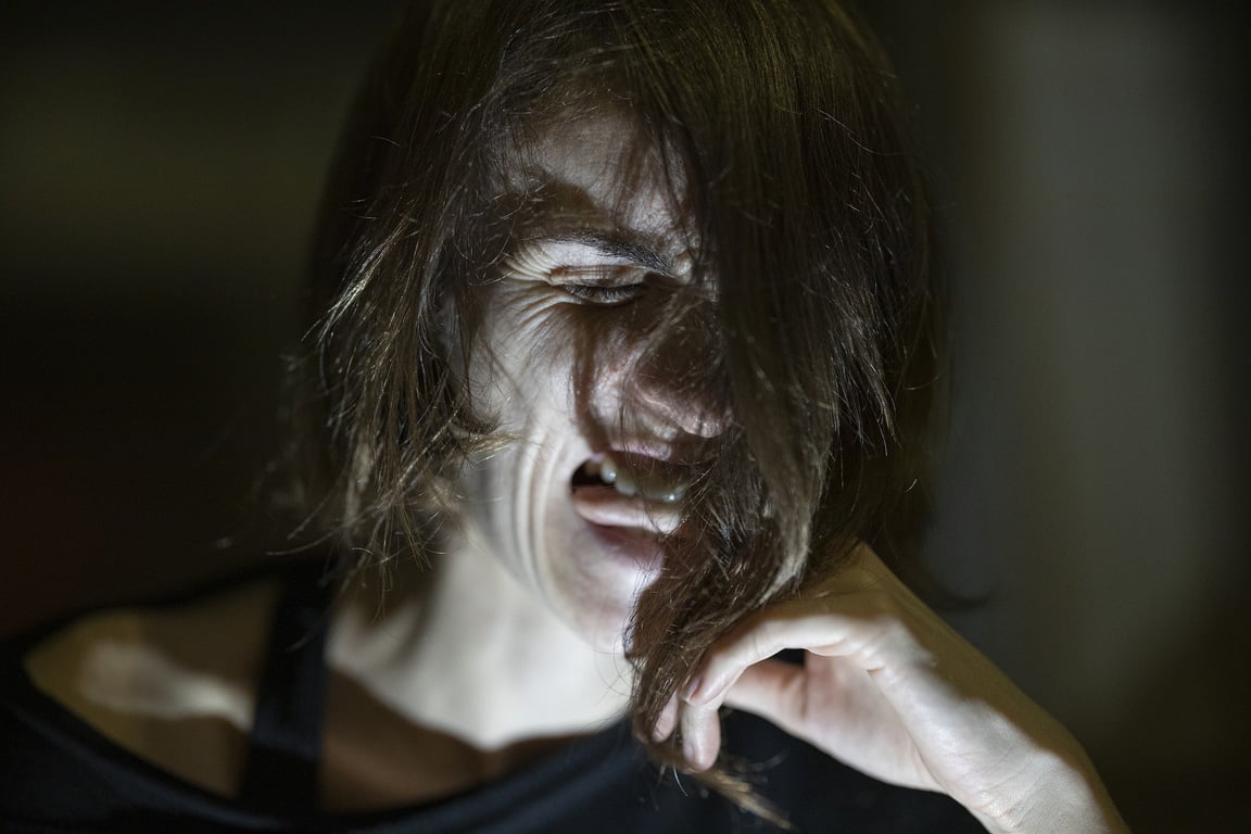 Fotografía de una mujer de cabello castaño, corto, cuyo rostro se encuentra iluminado por una luz blanca debajo de su barbilla, se encuentra con expresión de dolor, el resto de la fotografía tiene fondo negro.