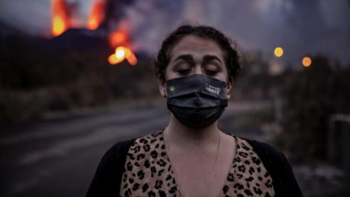 Fotografía de GN Diario que muestra a una mujer parada delante del escenario de un volcán en erupción. Ella es de edad madura, tiene discapacidad visual y los ojos cerrados, cabello oscuro recogido detrás de la cabeza, usa un cubre bocas negro justo bajo los ojos maquillados. Usa un vestido de cuello en V con estampado de leopardo y un suéter negro abierto.