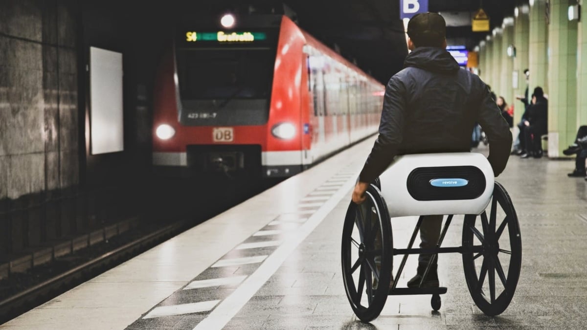 Fotografía de una estación de metro. En la parte izquierda se ve al tren color naranja llegando, mientras en el andén se ve de espaldas a un usuario de silla de ruedas, uno de los prototipos plegables. El usuario viste una chamarra negra con capucha que le cuelga sobre la espalda.