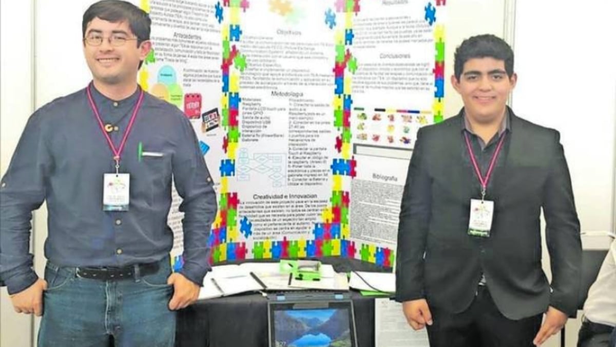 Fotografía de dos adolescentes varones que están de pie frente a un mosaico de información que tiene márgenes con rompecabezas de colores en alusión al trastorno del espectro autista y en medio de ellos hay un equipo de cómputo y documentos. Los dos jóvenes tienen colgando de su cuello una tarjeta de acreditación blanca con una cinta color vino. El primero de ellos usa lentes y viste una camisa de manga larga color gris y pantalón de mezclilla, el segundo viste de oscuro. Usa pantalón y saco cerrado negro con camisa negra.