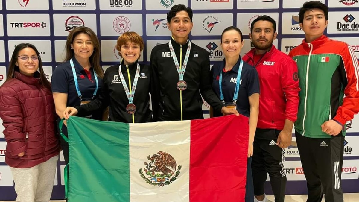 Fotografía de un grupo de siete personas, cuatro de ellas mujeres, que forman parte de la selección mexicana de para taewkondo. Al centro, vestidos con el uniforme oficial color negro está una mujer y un hombre con medallas al cuello y grandes sonrisas, ambos flanqueados por sus entrenadoras que también lucen sus medallas. Los dos deportistas que obtuvieron presea sostienen con sus manos una bandera mexicana desplegada.