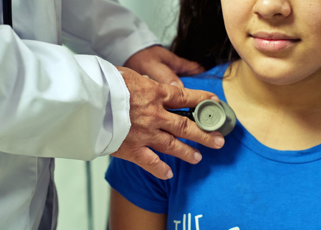 Fotografía que muestra las manos de un doctor que tiene piel gruesa y morena con dedos anchos y arrugados; y, se aprecia que viste bata blanca, se encuentra colocando un estetoscopio sobre el pecho de una niña de tez morena, cuyo rostro no se aprecia completamente, solo la nariz, ancha de la punta, labios carnosos y viste una blusa azul celeste, se alcanza a apreciar que tiene el cabello de color negro y largo.