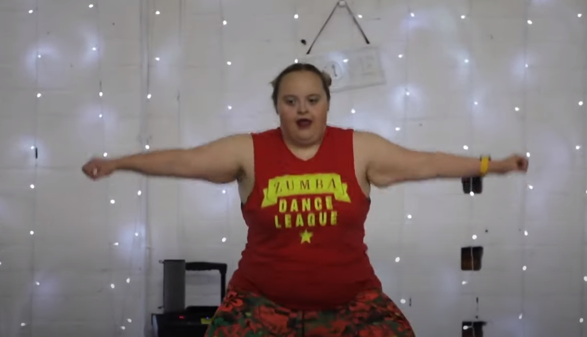 Captura de pantalla de Hannah Payton, una mujer joven, de tez blanca, cabello rubio, recogido en dos chongos, se encuentra frente a una pared blanca con luces, viste una playera de color rojo que tiene estampada en color amarillo la frase “Zumba Dance League”, ella se encuentra de pie, con los brazos extendidos a los lados y ambas piernas abiertas en compás, por la calidad de la imagen su rostro no luce a detalle, pero se percibe que tiene ojos pequeños de color café oscuro y se encuentra con la boca abierta.