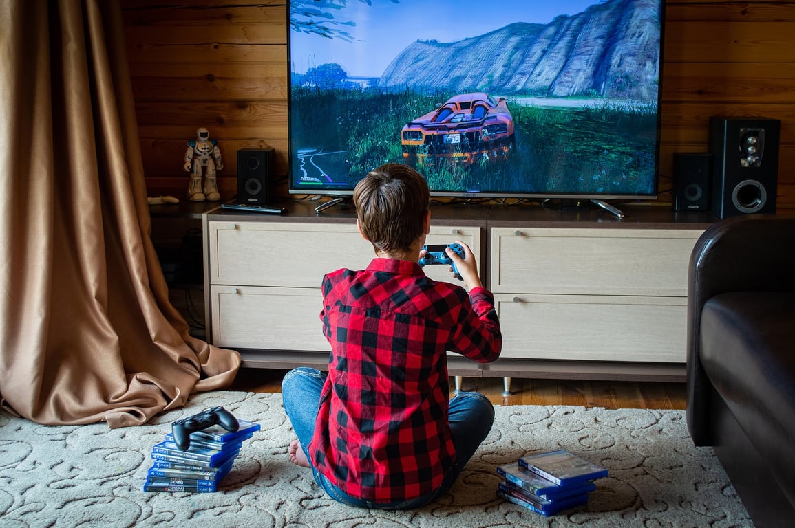 Fotografía de un niño de aproximadamente 12 años de espaldas a la cámara, viste una camisa a cuadros de color negro y rojo, se encuentra de frente al televisor que proyecta un automóvil de color naranja y tiene en ambas manos un control remoto de color azul, a su costado hay una pila de nueve CD’s de videojuegos de color azul.