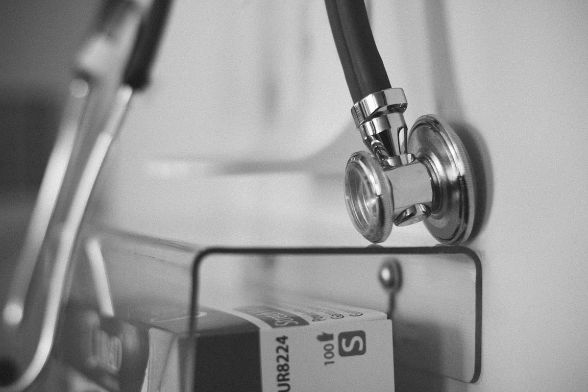 Fotografía a blanco y negro de un estetoscopio colgado en la pared, debajo del cual se encuentra un contenedor de vidrio en forma rectangular con un par de cajas de medicamento dentro.