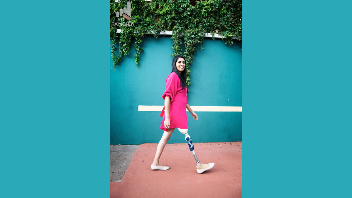 Fotografía de una mujer joven, de cabello castaño oscuro, largo a la altura del pecho, sonríe frente a la cámara, lleva puesto un vestido de color rosa mexicano y zapatos abiertos de color blanco, en su pierna izquierda tiene una prótesis de metal que hasta abajo tiene la forma de un pie. La mujer se encuentra caminando por la banqueta dde color naranja y detrás de ella hay una barda azul esmeralda con varias ramas de árbol de color verde oscuro.