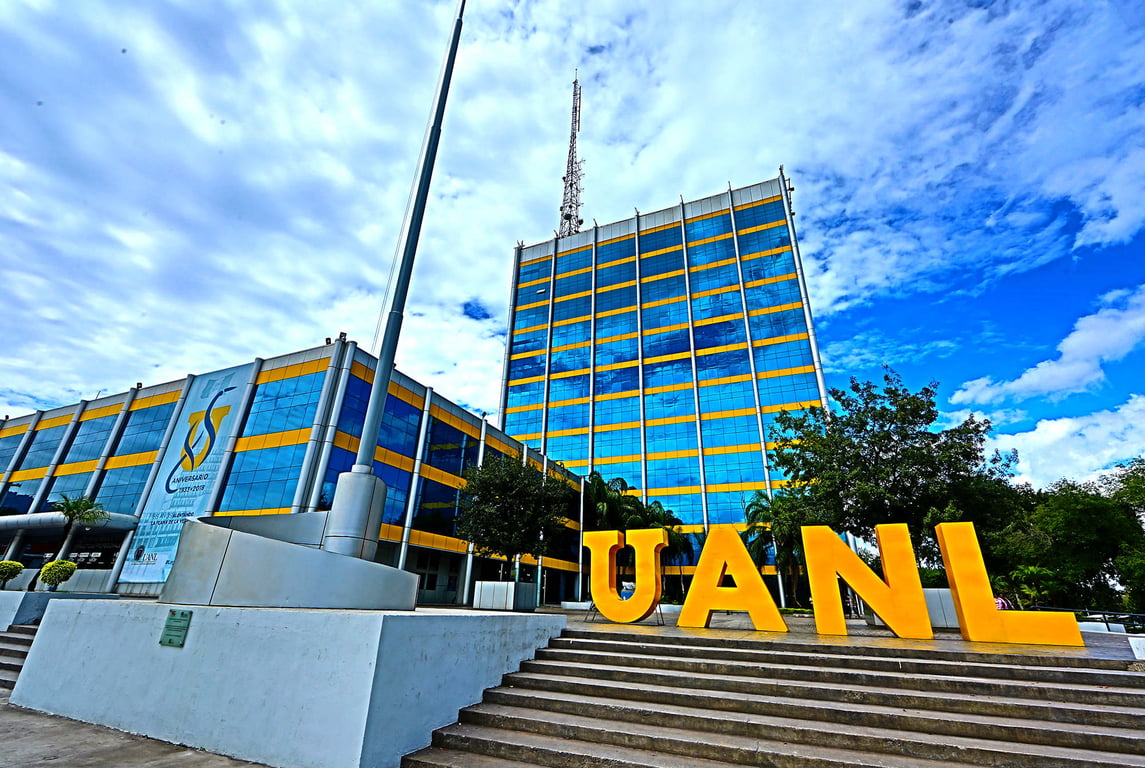 Fotografía de la entrada de la Universidad Autónoma de Nuevo León, un edificio alto con vidrios que reflejan el cielo, tiene unas escaleras de cemento con un letrero de color amarillo de letras en 3D que dicen UANL, el cielo detrás del edificio es de color azul con nubes de color blanco.