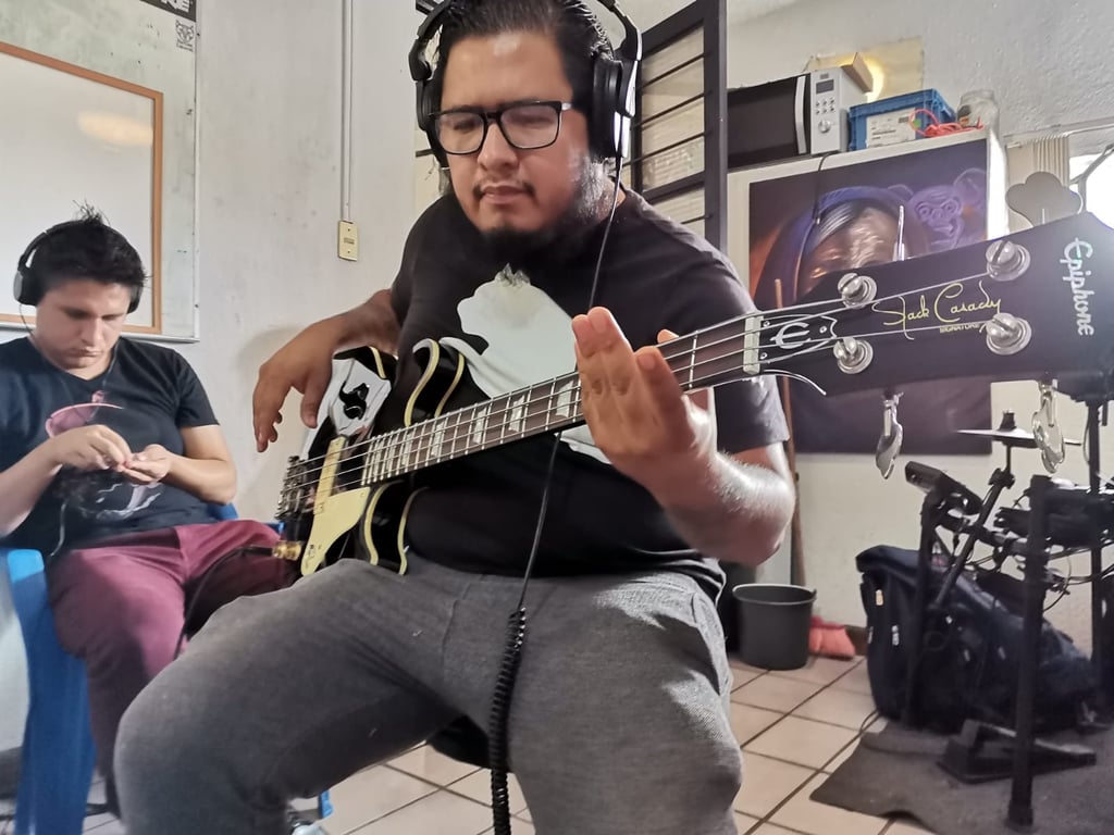 Fotografía de Andrés Otokani un hombre de edad media, tez morena claro, barba de color negro y tipo candado, utiliza anteojos cuyo armazón es de color negro, viste una playera negra con el logotipo de un puma al centro y un pants de color gris, se encuentra con unos audífonos de gran tamaño sobre su cabeza y toca con ambas manos una guitarra eléctrica de color negro con beige, mira al piso concentrado.