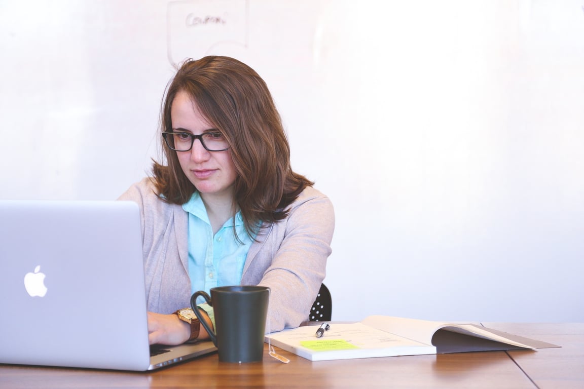 Fotografía de una mujer de edad media, cabello castaño, lentes cuyo armazón es de color negro, viste una blusa de color azul y un saco de color beige, se encuentra sentada en un escritorio detrás de una computadora portátil de la marca Apple.