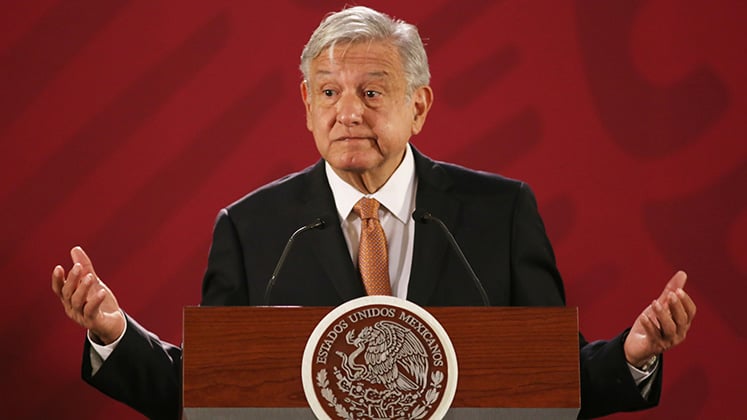 Fotografía del presidente Andrés Manuel López Obrador un hombre de edad adulta, cabello cano, rostro ovalado con expresión sonriente, está parado detrás de un atril con el escudo de México, hace un gesto torciendo la boca, viste un traje de color negro y corbata color naranja.