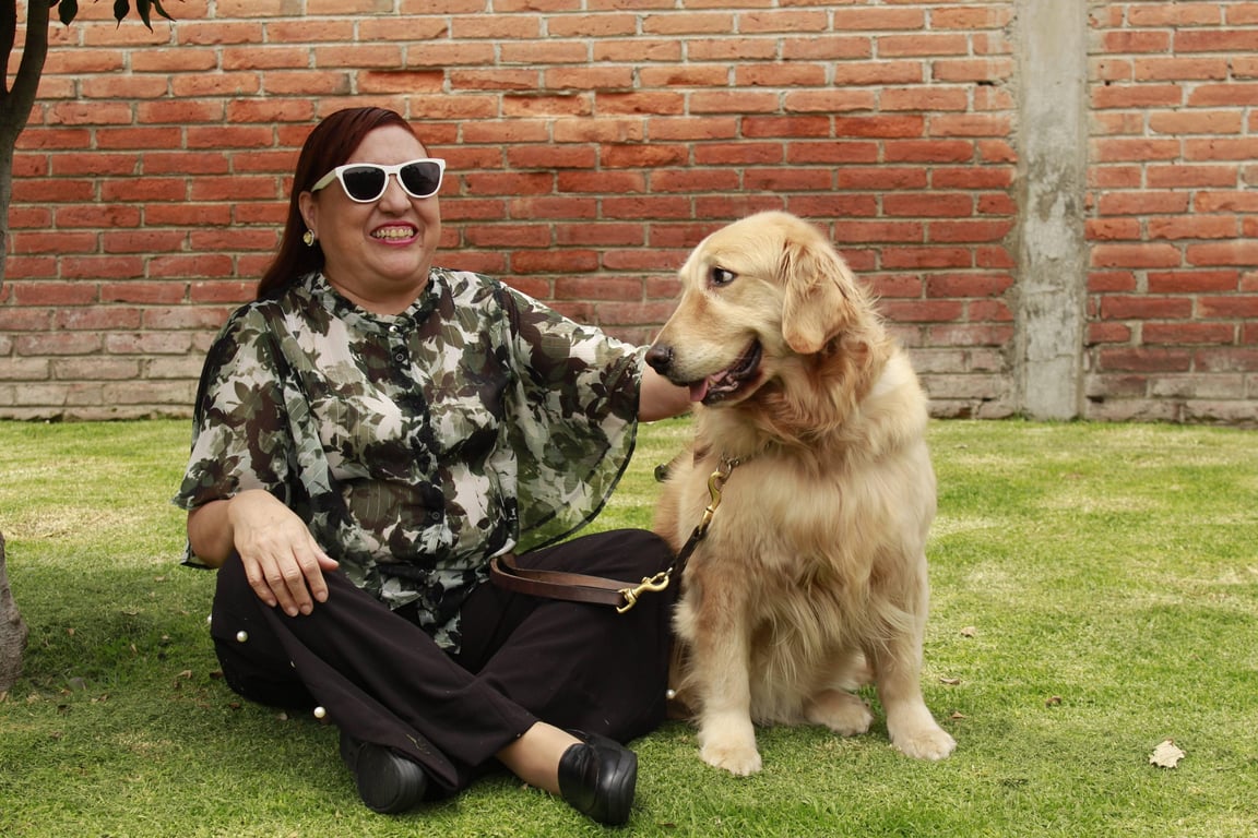 Fotografía a color de cuerpo completo de Silvia Lozada Badillo y su perra guía Kitty, Silvia es una mujer ciega de 59 años, mexicana, en las instalaciones de la escuela para entrenamiento de perros guía para ciegos, es un jardín con pasto verde y corto, un árbol y el fondo de una pared de ladrillos. Silvia se encuentra sentada en el pasto con las piernas cruzadas, su mano izquierda posa en su rodilla izquierda y con la otra acaricia a su perra guía, Kitty que se encuentra sentada y le dirige una mirada, que pareciera que le sonríe. Silvia es una mujer de piel morena, complexión mediana, cabello ondulado, peinado como una trenza de diadema y ondulado en las puntas. Sus ojos no se aprecian, ya que usa lentes obscuros con pasta gruesa, labios delgados y gran sonrisa. Usa una blusa con estampado de flores y hojas en tonos verdes con botones al frente y amplias mangas, también lleva puesto un pantalón negro y zapatos negros de piso.