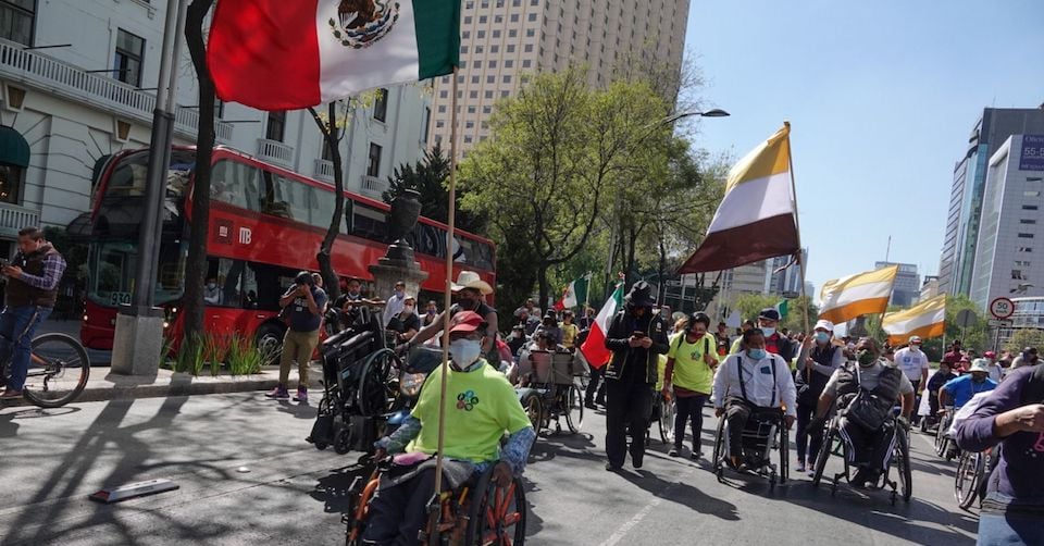 Usuario de silla de ruedas que sostiene una bandera de México en ambas manos se manifiesta sobre la avenida reforma.