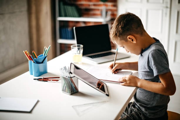Fotografía de un niño sentado en una silla frente a un escritorio de color blanco, se encuentra escribiendo en una libreta y tiene un tableta a su costado derecho, a su izquierda tiene una computadora portátil.