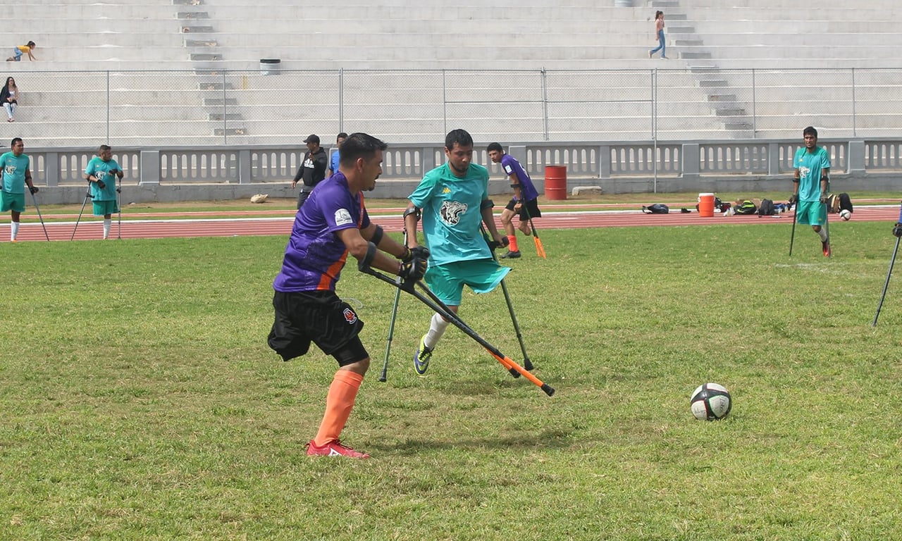 Dos jugadores dentro de una cancha de fútbol que tienen el pie izquierdo amputado peleando el balón con la muleta que los mantiene de pie.