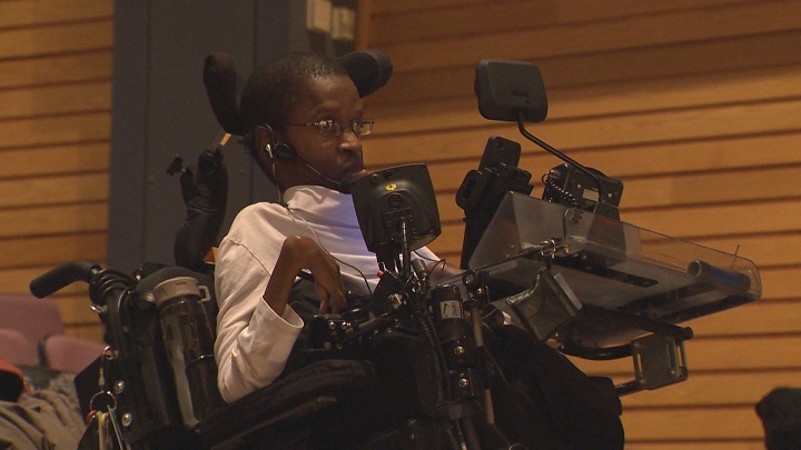 Gift Tshuma, usuario de silla de ruedas con aparente discapacidad motriz que tiene monitores y varios artículos tecnológicos frente a su rostro, sostenidos de un arnés adaptado a su silla.