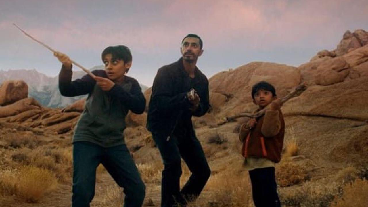 El actor Riz Ahmed de pie sobre una duna de arena con dos niños a su lado, ambos niños están en posición de defensa sosteniendo varas de árbol en sus manos.