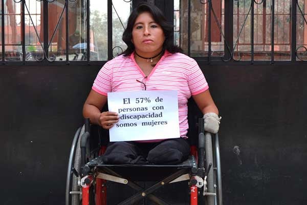 Mujer usuaria de silla de ruedas con una mano vendada y con la otra sostiene un letrero que dice "el 57 por ciento de las personas con discapacidad somos mujeres"