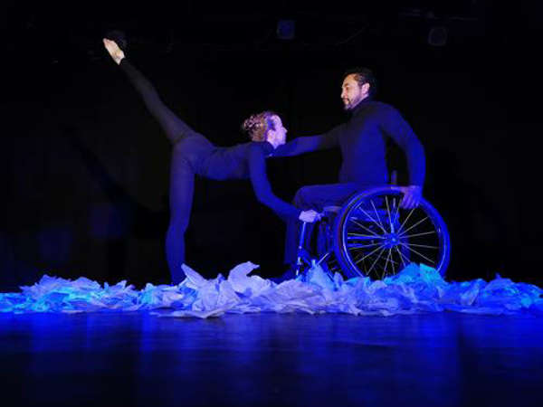 Dos personas danzando, un usuario de silla de ruedas y una mujer sostenida de su silla con un pie en el piso y otro al aire.