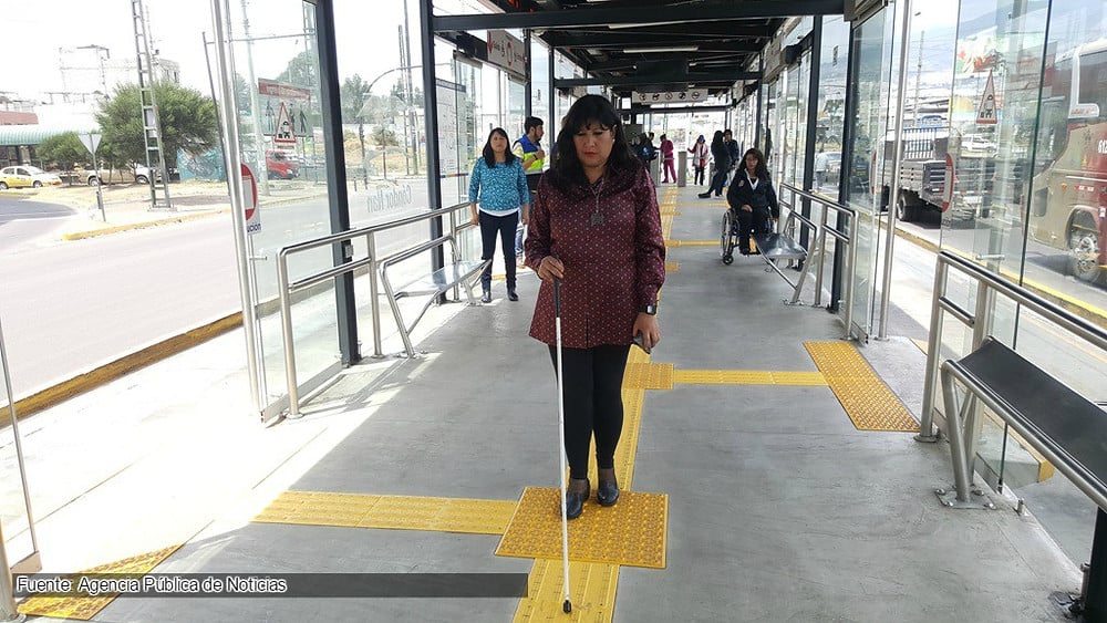 Mujer ciega de pie sobre las señales guía en el piso de una estación de transporte público.