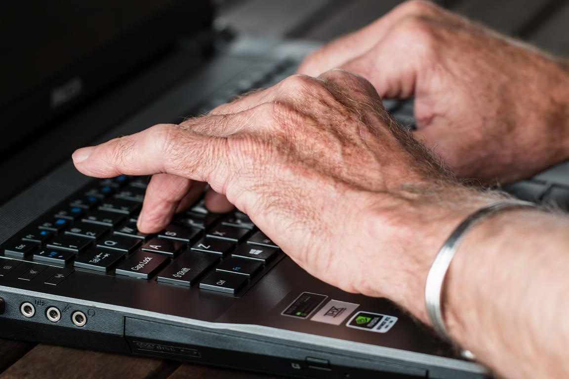 Manos arrugadas de adulto mayor tecleando letras en una computadora portátil.