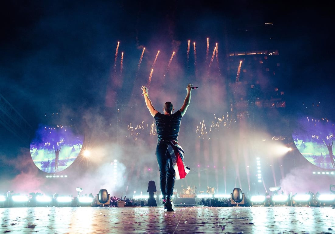 Vocalista de la banda Coldplay en el escenario con las manos extendidas hacia arriba.