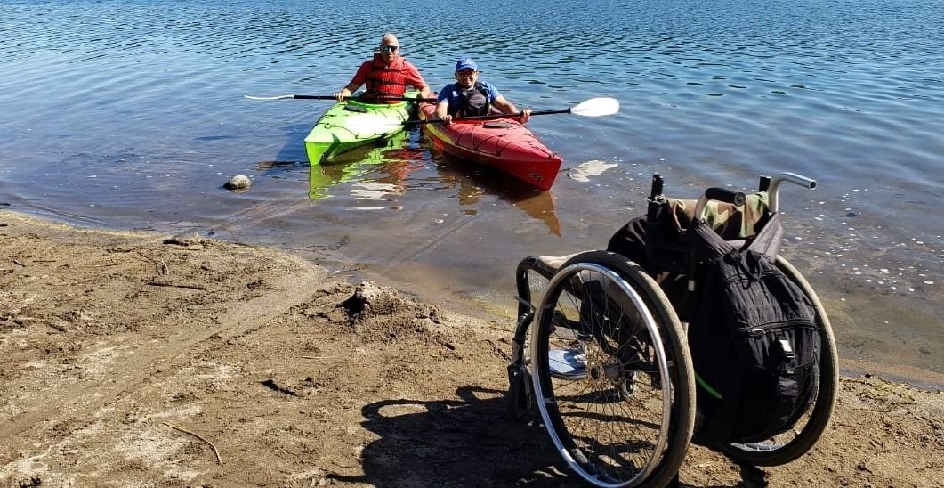 Silla de ruedas a la orilla del mar, su usuario se encuentra dentro del agua en una balsa de kayak.