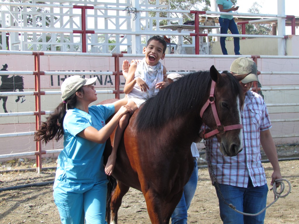 Niño con discapacidad motriz sobre un caballo apoyado por una instructora.
