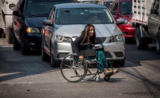 Mujer usuaria de silla de ruedas cruzando una avenida con carros transitando detrás de ella.