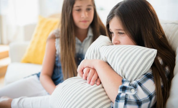 Dos niñas en una sala, una de ellas abraza una almohada de forma ansiosa y la otra niña intenta calmarla.