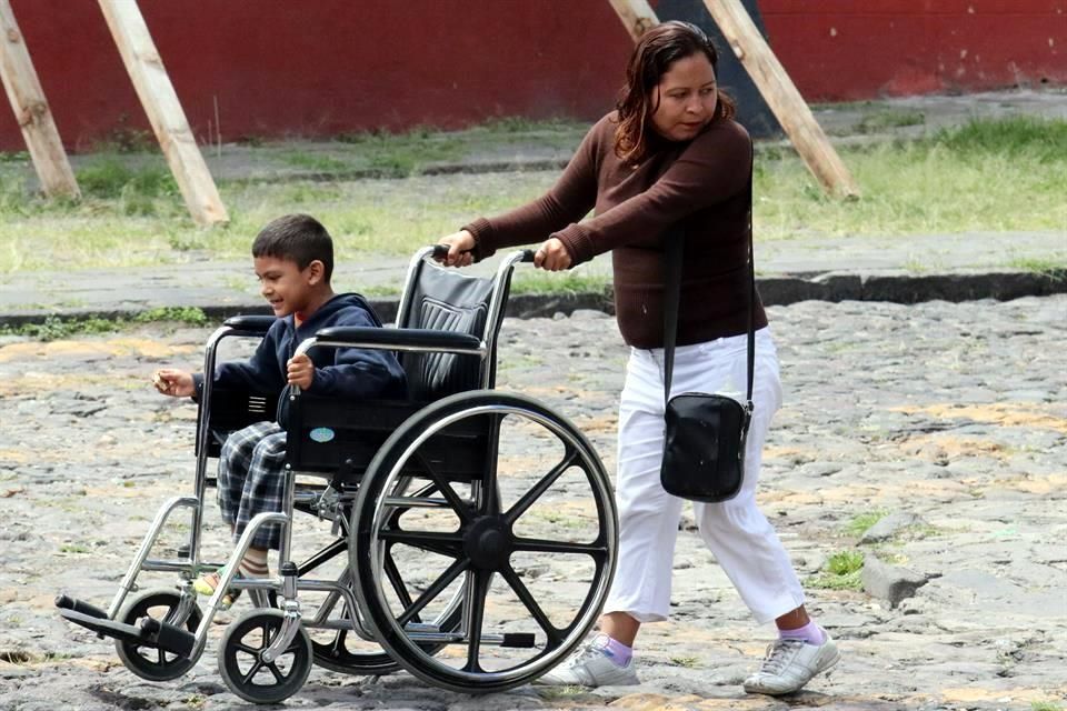 Mujer llevando con dificultad a su hijo usuario de silla de ruedas por una calle empedrada.