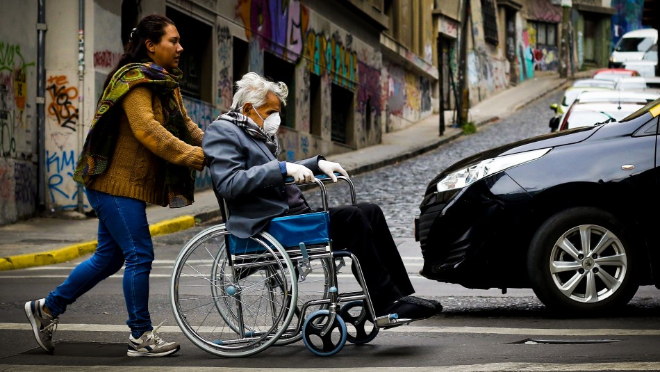 Cuidadora de un usuario en silla de ruedas empujándolo sobre la acera.